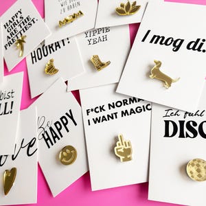 Könnte beinhalten: Eine Sammlung goldfarbener Metallanstecker auf weißen Präsentationskarten mit verschiedenen schwarzen Schriftzügen wie Happy Girls Are The Prettiest, I Mog Di, Yippie Yeah, F*ck Normal I Want Magic, Be Happy und Ich Fuhl Mich Disco.