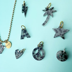 Könnte beinhalten: Goldfarbene Halskette mit Anhänger "HAPPY" und einem Buchstaben "M". Weitere Anhänger sind Blitz, Palme, Stern und Peace-Zeichen, alle mit grau-schwarzem Leopardenmuster. Verschiedene andere Anhänger sind ebenfalls sichtbar.