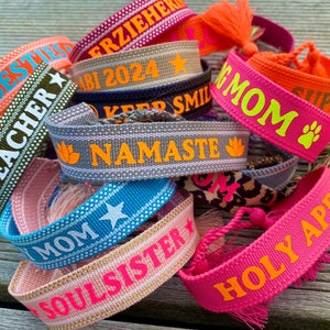 Könnte beinhalten: Eine Sammlung bunter gewebter Armbänder mit verschiedenen Sprüchen, darunter "Best Teacher", "Mom", "Namaste" und "Holy Apero".