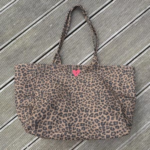 Könnte beinhalten: Große Tragetasche mit braunem und schwarzem Leopardenmuster. Die Tasche hat zwei geflochtene Griffe und ein kleines rosa herzförmiges Detail auf der Vorderseite. Die Tasche ist aus Stoff.