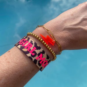 Può includere: Un braccialetto con stampa leopardata nera e rosa con il testo "GIRL MOM" in lettere rosa. Il braccialetto è indossato su un polso con un braccialetto a catena dorata e un ciondolo a forma di fulmine rosso.