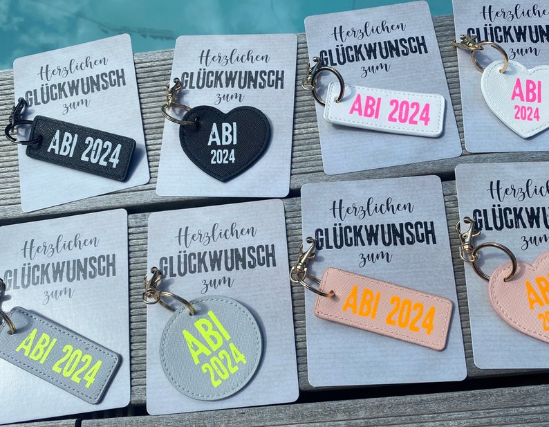 May include: A set of eight keychains with different colored leather and fabric designs. Each keychain features the text "Herzlichen Gl&uuml;ckwunsch zum ABI 2024" and a variety of shapes, including a heart, a rectangle, and a circle.