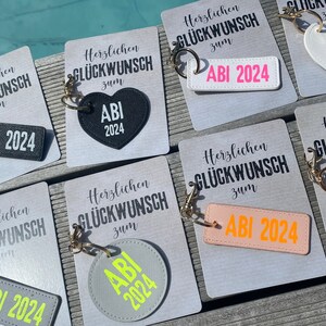 May include: A set of seven keychains with different colored heart and rectangular shapes. Each keychain has the text "Herzlichen Gl&uuml;ckwunsch zum ABI 2024" and a heart or rectangle with the text "ABI 2024" in a contrasting color.