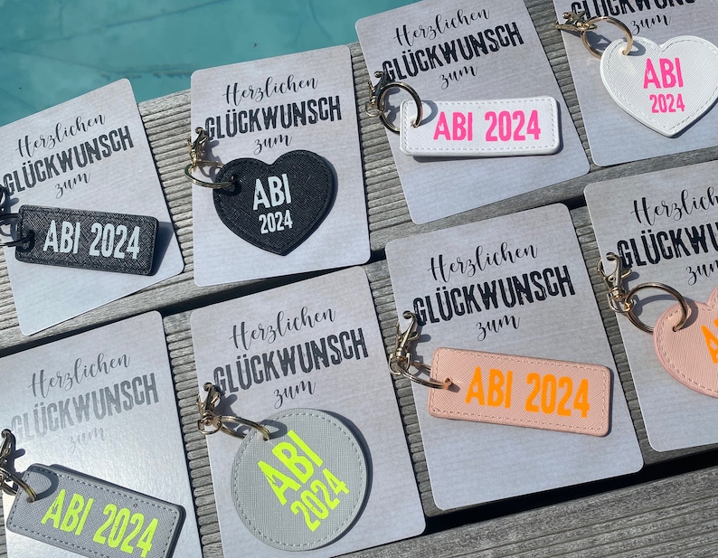 May include: A set of seven keychains with different colored heart and rectangular shapes. Each keychain has the text "Herzlichen Gl&uuml;ckwunsch zum ABI 2024" and a heart or rectangle with the text "ABI 2024" in a contrasting color.