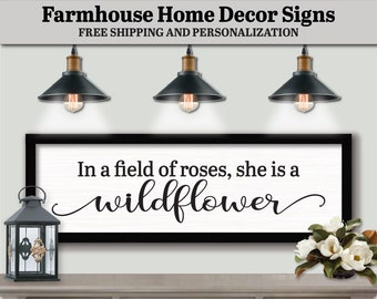 May include: Black and white farmhouse style sign with the text "In a field of roses, she is a wildflower". The sign is hanging on a wall above a gray wooden dresser with two drawers. There are three black pendant lights hanging above the sign.