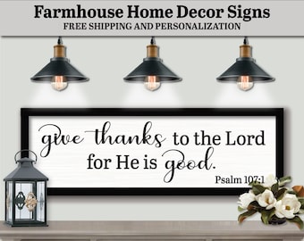 May include: Black and white farmhouse style sign with the text "give thanks to the Lord for He is good. Psalm 107:1". The sign is hanging on a wall with a black frame and is displayed above a gray console table.