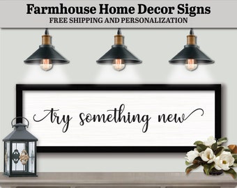 May include: Black and white farmhouse sign with the text "try something new" in a cursive font. The sign is framed in black and hanging on a wall with three black pendant lights above it.