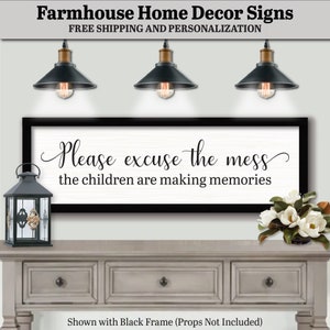 May include: White farmhouse sign with black text that reads "Please excuse the mess the children are making memories". The sign is framed in black and hanging on a wall above a gray dresser with two drawers. There are three black pendant lights hanging above the sign.