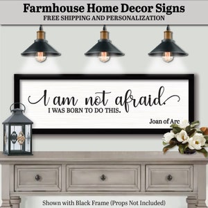 May include: A farmhouse-style sign with a black frame and the quote "I am not afraid, I was born to do this." The sign is displayed on a gray console table, with a decorative lantern and a bouquet of white flowers.
