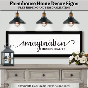 May include: A farmhouse style sign with a black frame, featuring the text "imagination creates reality" in a decorative script font. The sign is displayed on a whitewashed wood surface with a decorative lantern and a vase of white flowers.