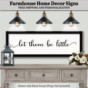 May include: Black framed farmhouse sign with the text "let them be little" hanging above a white dresser with two drawers. The sign is displayed with three black pendant lights and a black lantern.