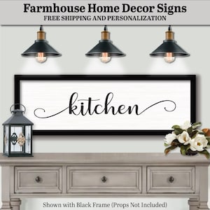 May include: Black and white farmhouse style sign with the word "kitchen" in a cursive font. The sign is framed in black and hanging on a wall above a gray wooden dresser with two drawers.
