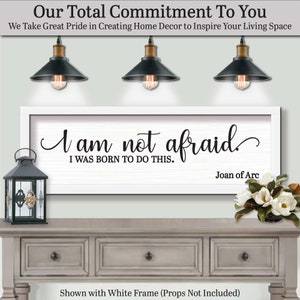 以下が含まれることがあります： 「I am not afraid, I was born to do this」の引用句が書かれた、白いフレームのウォールアート。ジョーン・オブ・アークによるもの。グレーのコンソールテーブルの上に飾られており、黒いランタンとフラワーアレンジメントが添えられています。