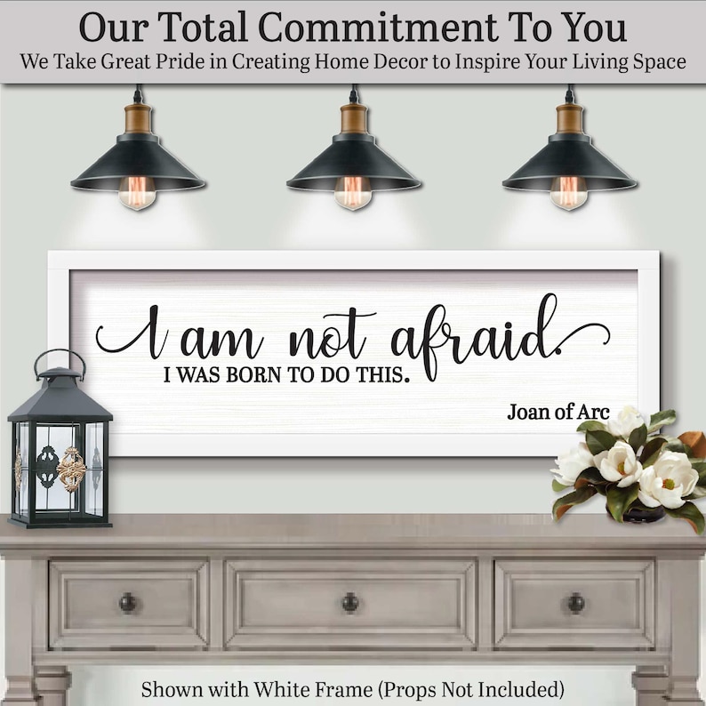 以下が含まれることがあります： 「I am not afraid, I was born to do this」の引用句が書かれた、白いフレームのウォールアート。ジョーン・オブ・アークによるもの。グレーのコンソールテーブルの上に飾られており、黒いランタンとフラワーアレンジメントが添えられています。