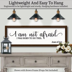 以下が含まれることがあります： 「I am not afraid, I was born to do this」というジャンヌ・ダルクの引用句が書かれた額入りの壁掛けアート。テキストは黒い筆記体で白い背景に書かれ、茶色のフレームで囲まれています。画像には、装飾的なランタン、花、3つのペンダントライトも表示されています。