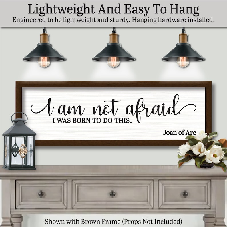 以下が含まれることがあります： 「I am not afraid, I was born to do this」というジャンヌ・ダルクの引用句が書かれた額入りの壁掛けアート。テキストは黒い筆記体で白い背景に書かれ、茶色のフレームで囲まれています。画像には、装飾的なランタン、花、3つのペンダントライトも表示されています。