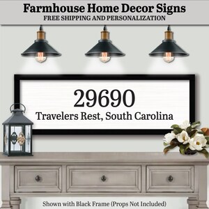 May include: A white farmhouse sign with black text that reads "29690 Travelers Rest, South Carolina". The sign is framed in black and is hanging on a wall above a gray wooden dresser. Three black pendant lights hang above the sign.