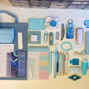 May include: A collection of blue stationery items including a tote bag, a quilted pouch, a daily planner, a reading log, bookmarks, pens, a notebook with a heart, a beaded bracelet, a feather, a keychain, and various office supplies arranged on a surface.