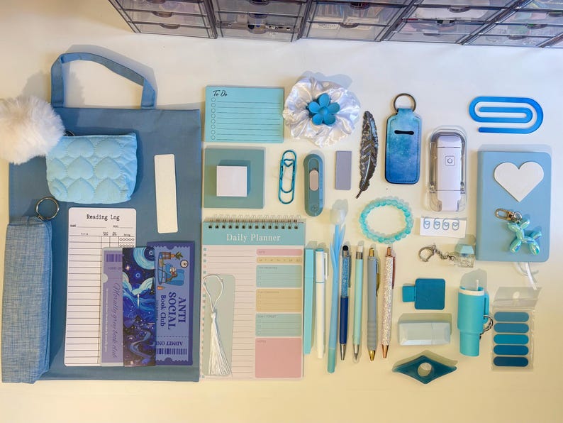 May include: A collection of blue stationery items including a tote bag, a quilted pouch, a daily planner, a reading log, bookmarks, pens, a notebook with a heart, a beaded bracelet, a feather, a keychain, and various office supplies arranged on a surface.