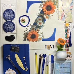 May include: A collection of stationery and accessories including a floral letter Z tote bag, a blue notebook with a sunflower sticker, various pens, a small potted crochet sunflower, sticky notes, car coasters, bookmarks, and a small black quilted bag.