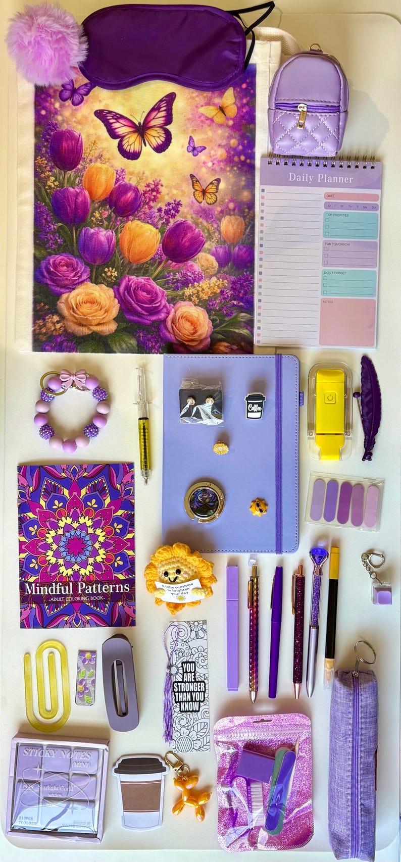 May include: A collection of purple and yellow stationery items including a floral notebook, a daily planner, a Mindful Patterns coloring book, a purple journal, pens, a pencil case, a crochet sunflower, a sleep mask, and various small accessories.