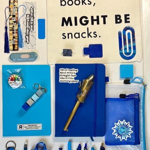 May include: A collection of blue-themed stationery and accessories including a canvas tote bag with text, a clipboard, various pens, a notebook with a sticker, a small plush shark, a blue sleep mask, bookmarks, and a clear zippered pouch.