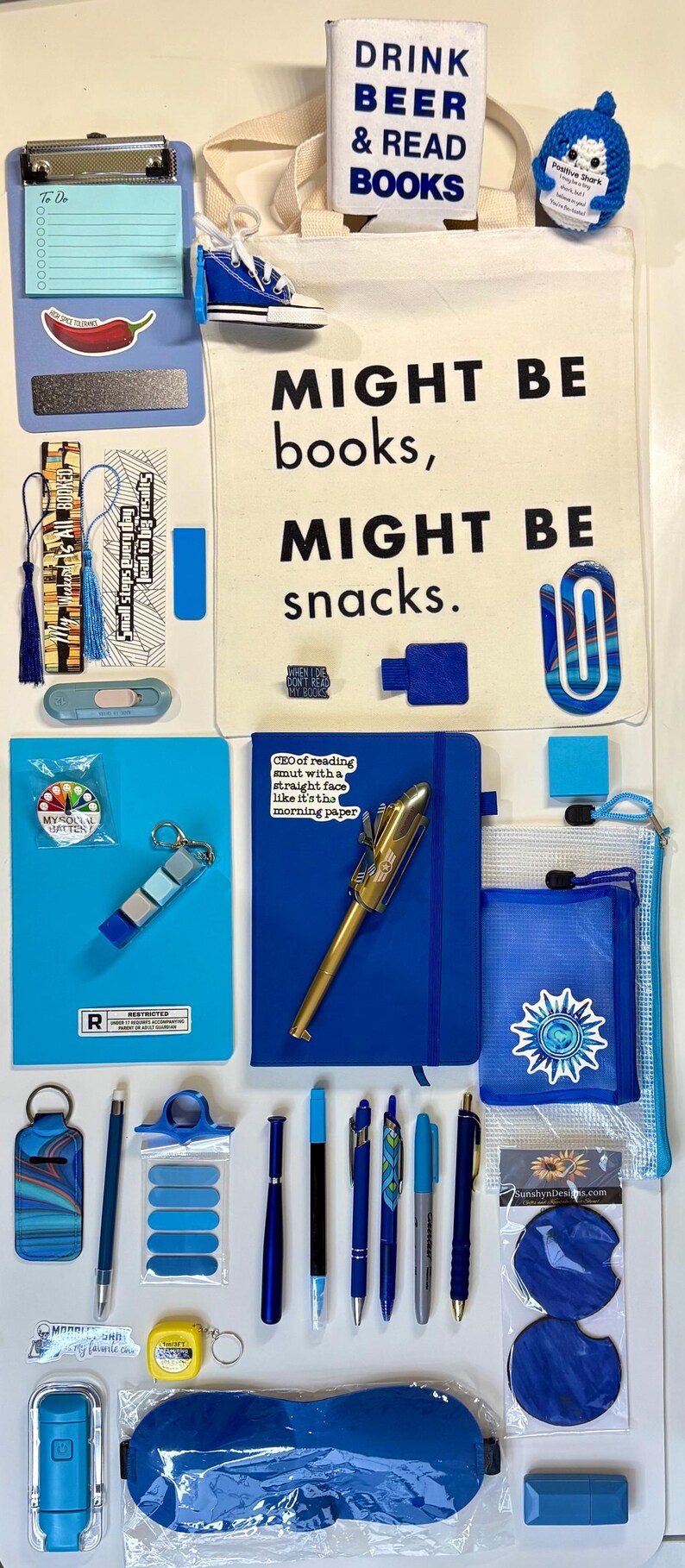 May include: A collection of blue-themed stationery and accessories including a canvas tote bag with text, a clipboard, various pens, a notebook with a sticker, a small plush shark, a blue sleep mask, bookmarks, and a clear zippered pouch.