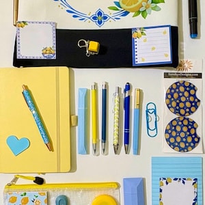 May include: A collection of stationery items including a white tote bag with a blue letter E and lemon motif, a yellow notebook, various pens, sticky notes with lemon patterns, a daily planner, a pencil case, and two circular car coasters.