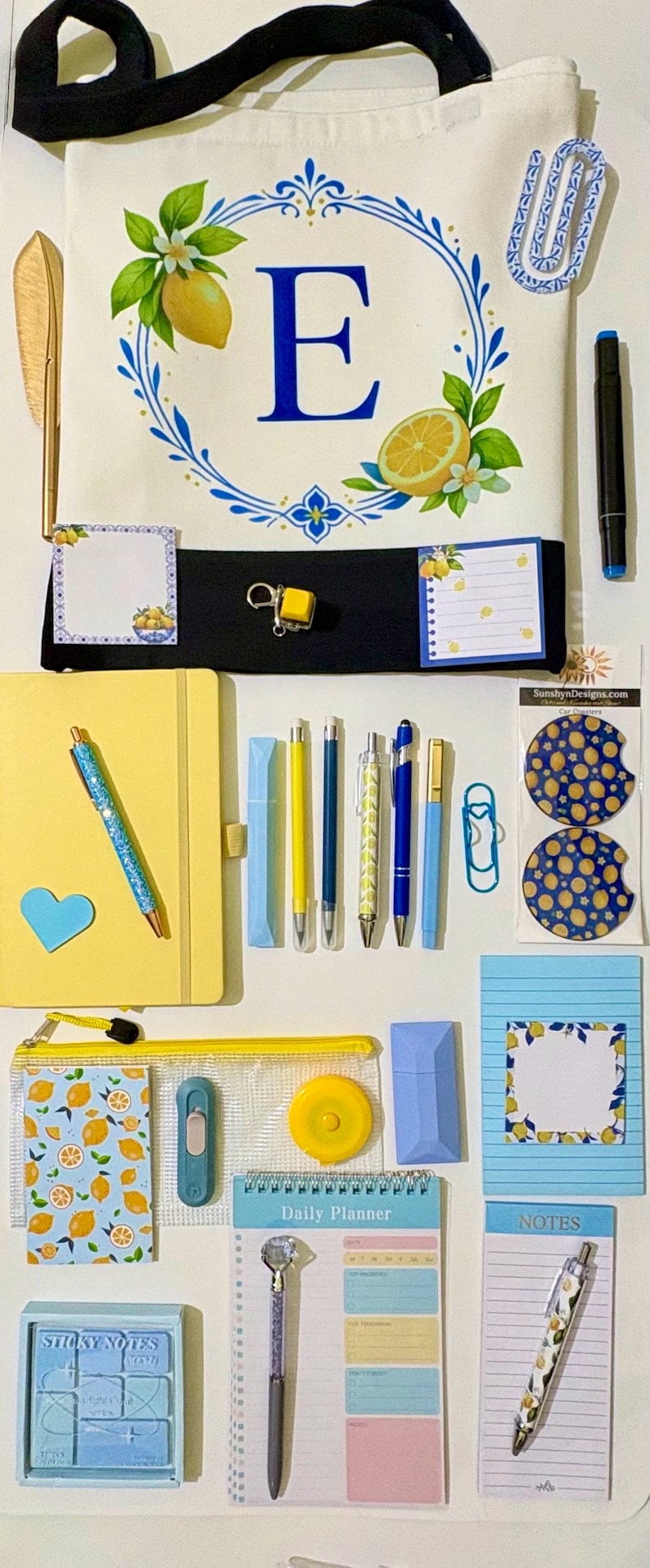 May include: A collection of stationery items including a white tote bag with a blue letter E and lemon motif, a yellow notebook, various pens, sticky notes with lemon patterns, a daily planner, a pencil case, and two circular car coasters.