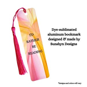May include: A rectangular aluminum bookmark with a dye-sublimated abstract pattern in shades of pink, orange, and yellow. The text "I'D RATHER BE READING" is printed in black. A bright pink tassel is attached to the top through a punched hole.