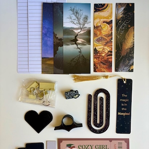 May include: A collection of reading accessories including two white reading log cards, a metal feather bookmark, four vertical bookmarks with scenic and artistic prints, gold binder clips, a book-shaped pin, a black heart, a black page holder, a large paperclip, a black bookmark with text, and a Cozy Girl Book Club ticket.