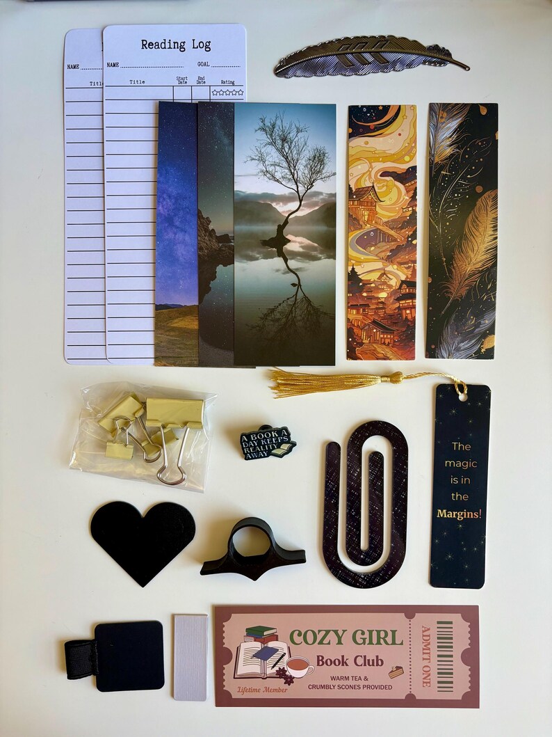 May include: A collection of reading accessories including two white reading log cards, a metal feather bookmark, four vertical bookmarks with scenic and artistic prints, gold binder clips, a book-shaped pin, a black heart, a black page holder, a large paperclip, a black bookmark with text, and a Cozy Girl Book Club ticket.