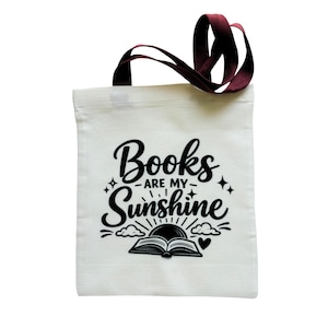 May include: A white canvas tote bag with two dark maroon handles. The front features black text that reads Books are my Sunshine, accompanied by a graphic illustration of an open book, sun rays, small clouds, a heart, and star accents.