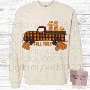 May include: A cream-colored crewneck sweatshirt featuring a graphic of a brown plaid pickup truck carrying pumpkins and pies. The text FALL VIBES is printed in orange below the truck. Orange pumpkins are positioned at the front and back wheels.
