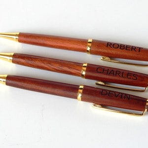 May include: Three personalized wooden pens with gold-colored accents. Each pen is engraved with a name: Robert, Charles, and Devin. The pens have a rich, dark brown color and a sleek, professional design.