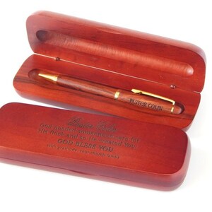 May include: A wooden pen and case set. The pen is brown with gold accents and the text "PASTOR COLIN". The case is open, revealing the pen, and has engraved text. The case is a reddish-brown color.