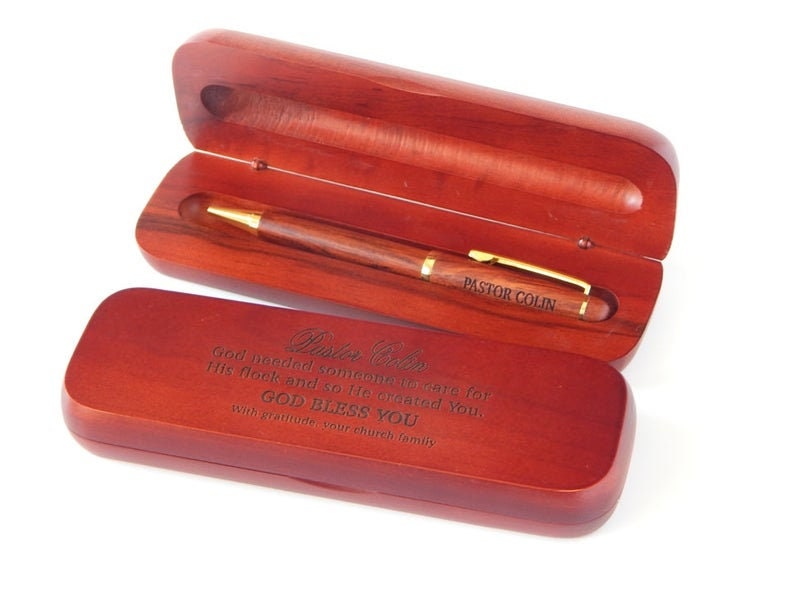May include: A wooden pen and case set. The pen is brown with gold accents and the text "PASTOR COLIN". The case is open, revealing the pen, and has engraved text. The case is a reddish-brown color.