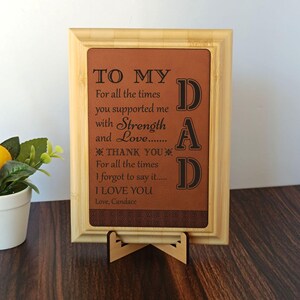 May include: A framed brown plaque with the text "TO MY DAD" and a heartfelt message. The frame is light brown, and the plaque is supported by a wooden stand. The message includes the words "Strength" and "Love."