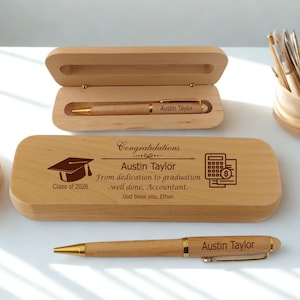 Op de afbeelding: Een houten pennen set met een pen en een etui. De etui is gegraveerd met de naam "Austin Taylor", een afstudeerhoed, een rekenmachine en de tekst "Class of 2026". De pen is ook gegraveerd met de naam "Austin Taylor".
