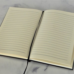 K&ouml;nnte beinhalten: Ein schwarzes, liniertes Notizbuch mit einem schwarzen Band als Lesezeichen. Das Notizbuch ist auf einer leeren Seite mit liniertem Papier ge&ouml;ffnet.