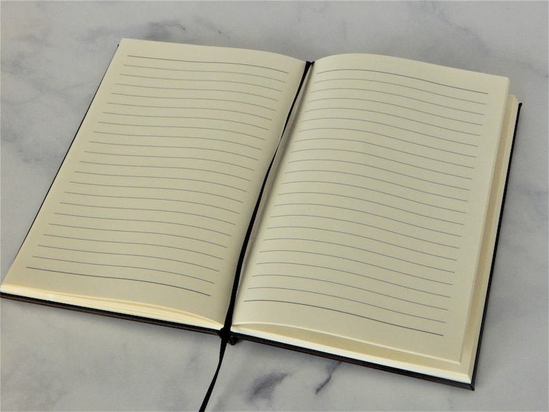 K&ouml;nnte beinhalten: Ein schwarzes, liniertes Notizbuch mit einem schwarzen Band als Lesezeichen. Das Notizbuch ist auf einer leeren Seite mit liniertem Papier ge&ouml;ffnet.