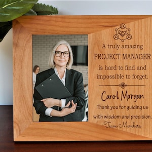 May include: A wooden picture frame with a photo of a woman in a business suit. The frame has engraved text that reads "A truly amazing PROJECT MANAGER is hard to find and impossible to forget. Carol Morgan. Thank you for guiding us with wisdom and precision."