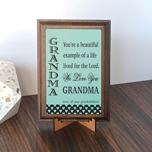 May include: A wooden framed sign with a light teal background and the word "GRANDMA" in large, black, capital letters. The sign includes the text "You're a beautiful example of a life lived for the Lord. We Love You GRANDMA. Love, all your grandchildren!"