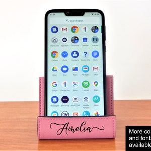 May include: A pink leather phone stand with the name Amelia engraved on it. The stand is holding a smartphone with a variety of apps on the screen.