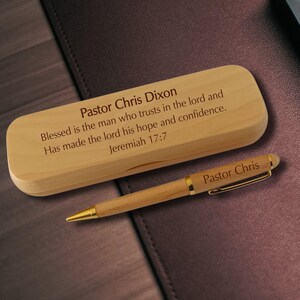 May include: A light-colored wooden pen case and a matching wooden ballpoint pen with gold-tone accents. The case is engraved with "Pastor Chris Dixon" and "Blessed is the man who trusts in the lord and Has made the lord his hope and confidence. Jeremiah 17:7". The pen is engraved with "Pastor Chris".