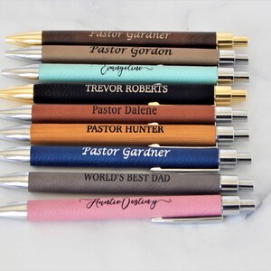 K&ouml;nnte beinhalten: Zehn personalisierte Stifte mit verschiedenen Leder- und Holz-Oberfl&auml;chen in verschiedenen Farben. Die Stifte sind mit Namen und Titeln graviert, darunter "Pastor Gardner", "Pastor Gordon", "Evangeline", "Trevor Roberts", "Pastor Dalene", "Pastor Hunter", "World's Best Dad" und "Auntie Destiny".