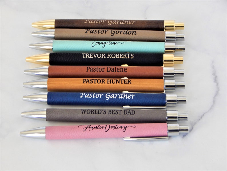 K&ouml;nnte beinhalten: Zehn personalisierte Stifte mit verschiedenen Leder- und Holz-Oberfl&auml;chen in verschiedenen Farben. Die Stifte sind mit Namen und Titeln graviert, darunter "Pastor Gardner", "Pastor Gordon", "Evangeline", "Trevor Roberts", "Pastor Dalene", "Pastor Hunter", "World's Best Dad" und "Auntie Destiny".