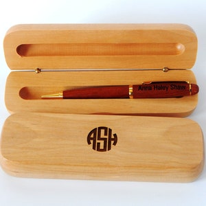 May include: A wooden pen and case set. The pen is a dark reddish-brown with gold accents and the inscription "Anna Haley Shaw." The light-colored wooden case is open, revealing the pen. The case has the initials "ASH" engraved on the lid.