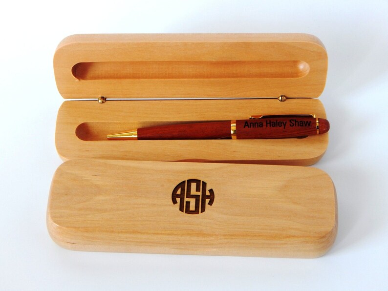 May include: A wooden pen and case set. The pen is a dark reddish-brown with gold accents and the inscription "Anna Haley Shaw." The light-colored wooden case is open, revealing the pen. The case has the initials "ASH" engraved on the lid.