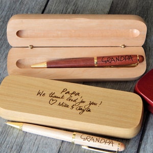 Peut inclure: Deux boîtes à stylos en bois avec des messages gravés. La boîte du haut contient un stylo marron avec des accents dorés et l'inscription "GRANDPA". La boîte du bas contient un stylo brun clair avec des accents dorés et l'inscription "GRANDPA". La boîte du bas contient également le message gravé "Papa, We thank God for you! Millie & Tayla".